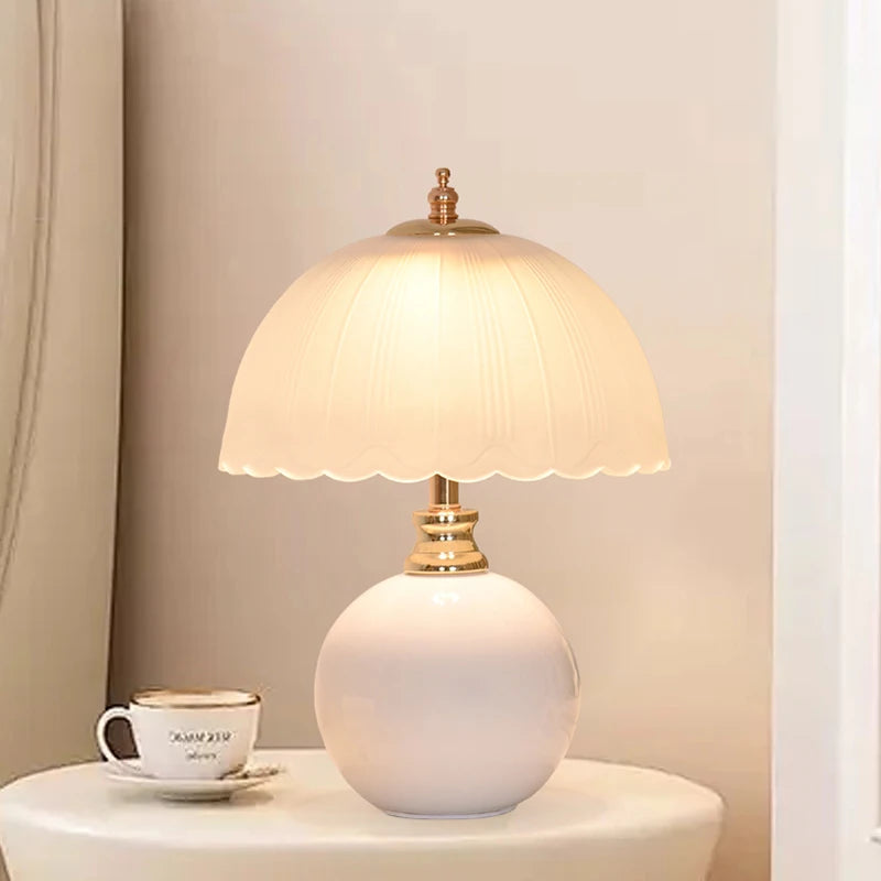 White ceramic-style table lamp with a scalloped shade glowing warmly on a bedside table in a calm neutral interior.